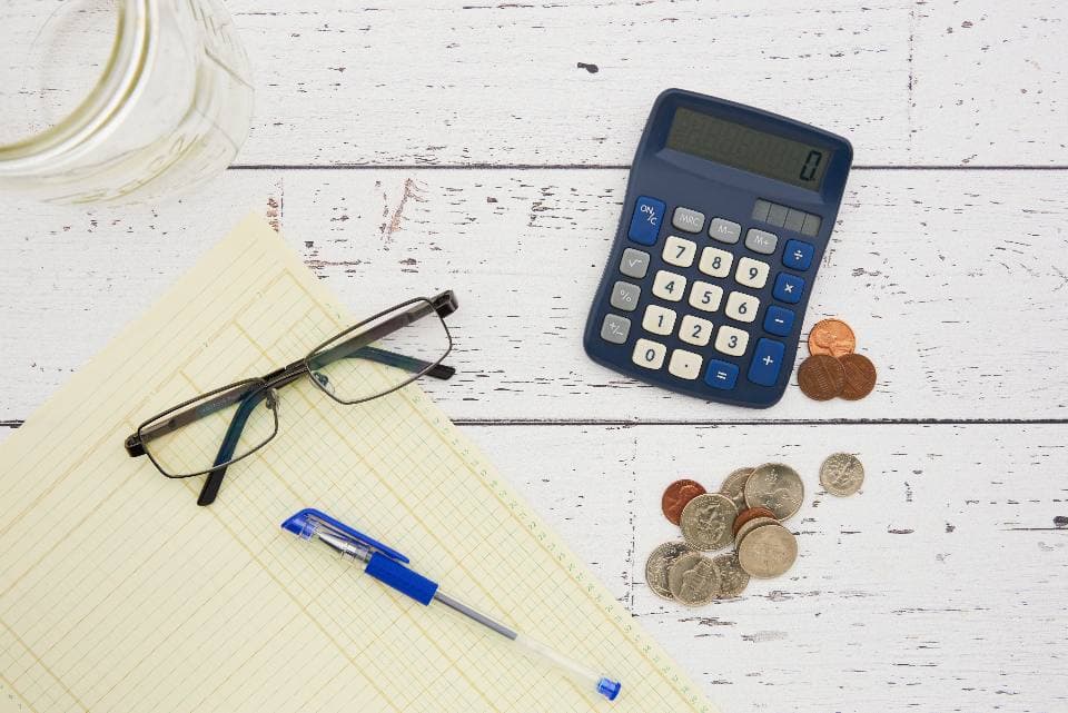 Overhead view of a blue desk calculator, reading glasses on a yellow ledger pad, a pen and small piles of coins on white painted wood.
