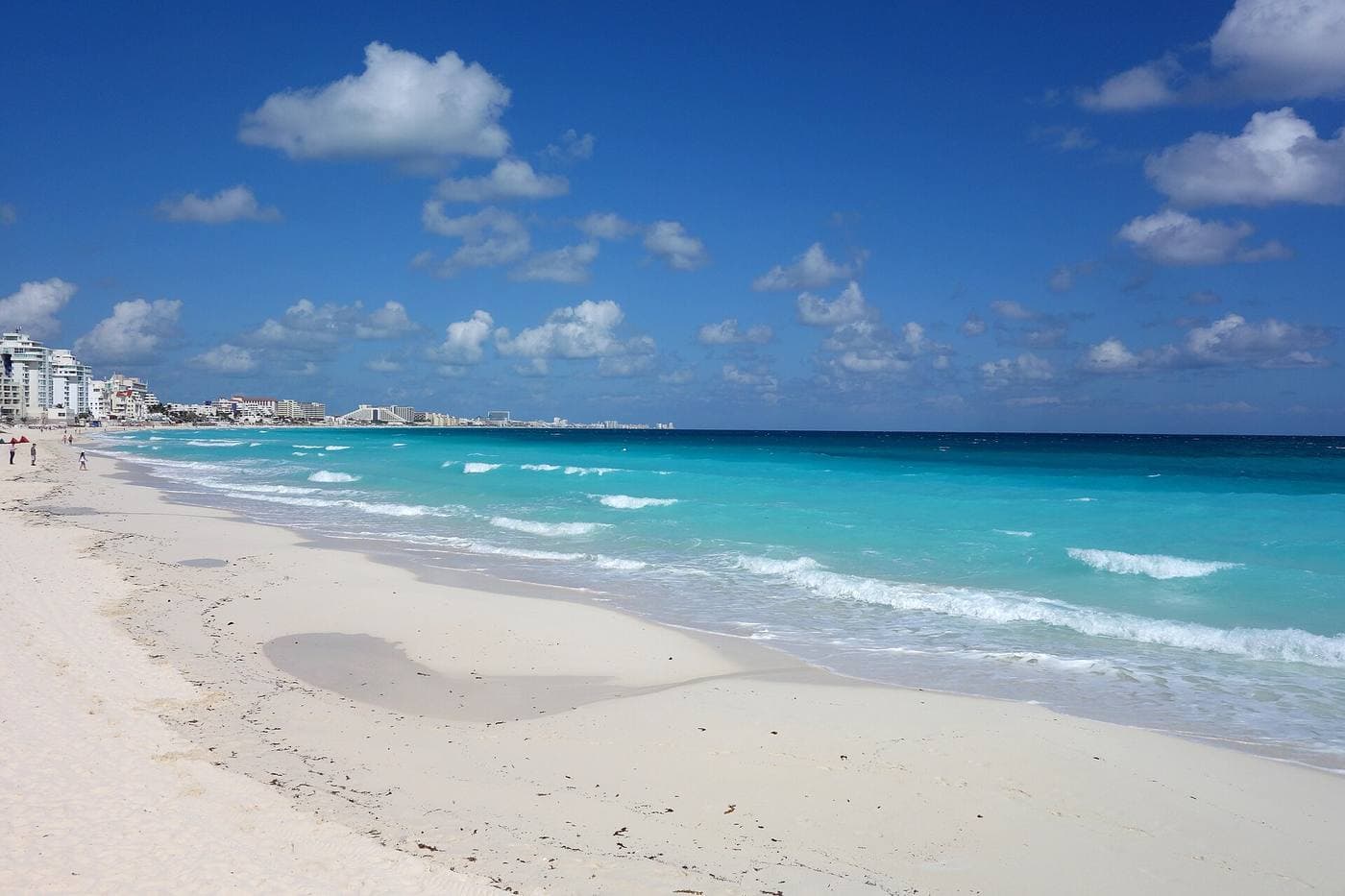 A broad curve of white sand meeting calm turquoise Caribbean water, resort towers in the far distance.
