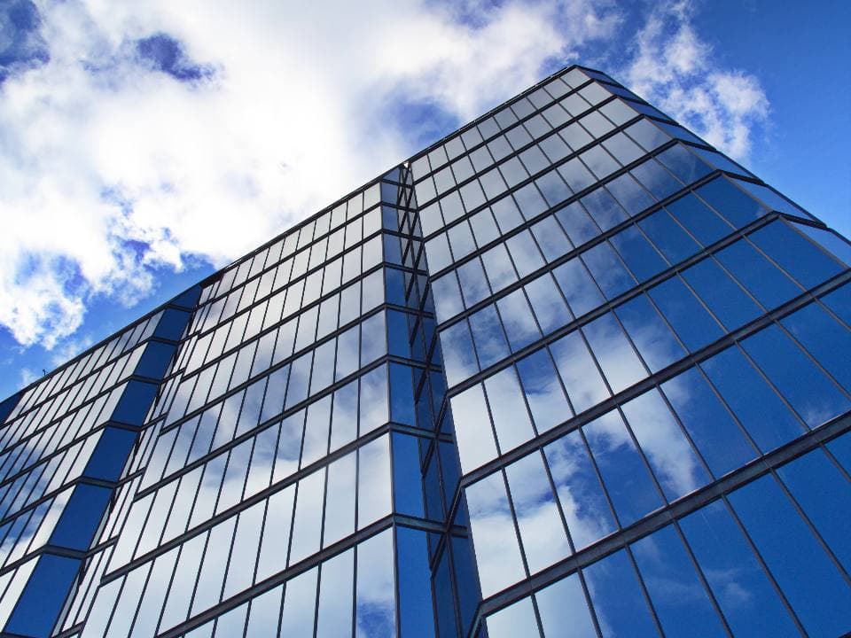 Low-angle view of a glass office tower whose windows mirror white clouds against a bright blue sky.