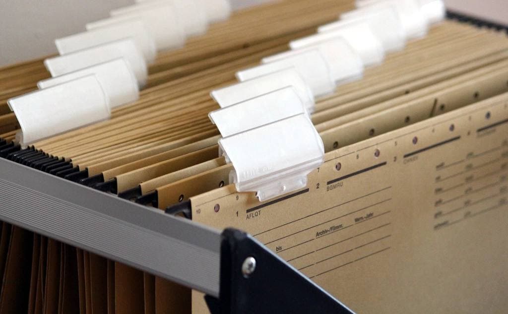 Rows of tan hanging file folders with blank white label tabs fill an open metal filing drawer, lit softly from above.