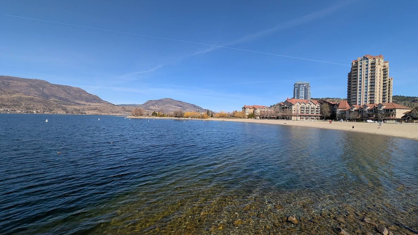 Clear Okanagan Lake water meeting a sandy beach below tall waterfront condo towers and red-roofed resort buildings, with dry brown hills across the lake