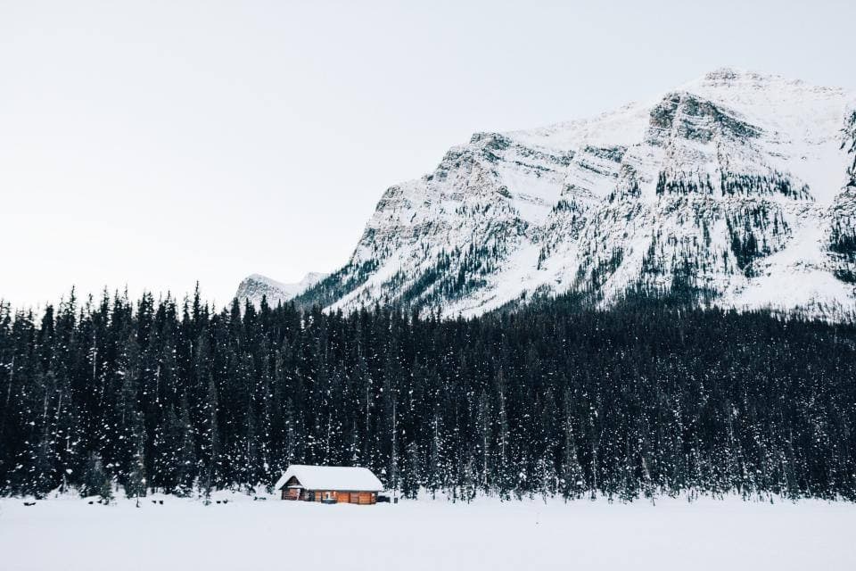 A small log cabin sits on a frozen snowfield at the edge of a dense evergreen forest, with a towering snow-covered mountain face behind.