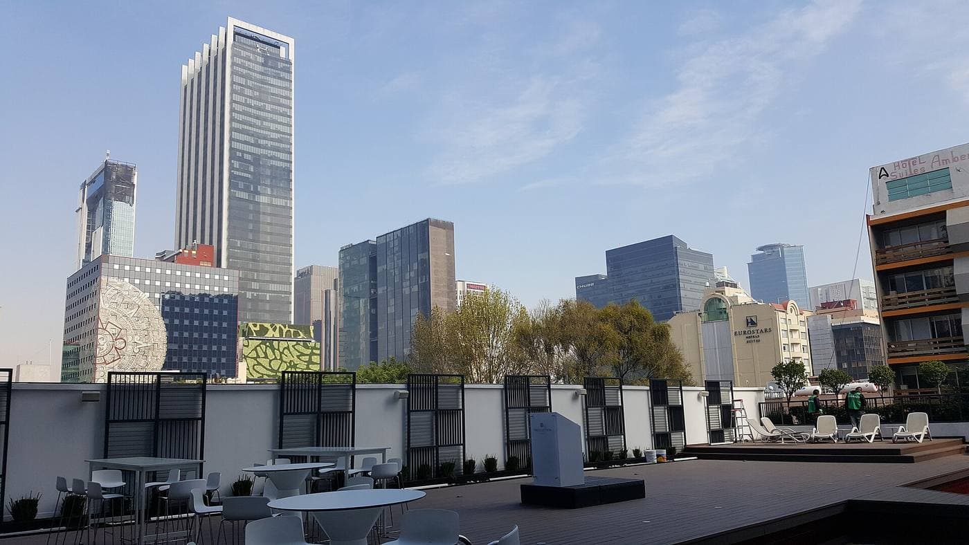 High-rise office and hotel towers of the Reforma corridor under a clear blue sky, seen across a rooftop terrace set with white tables and loungers.