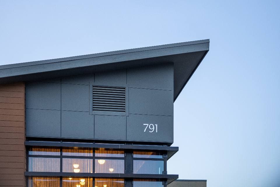 A modern grey-clad building corner with a cantilevered roof and the number 791, warm light glowing through its tall windows at dusk.