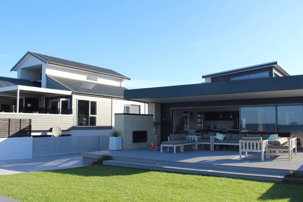 A contemporary home with dark cladding and sliding glass walls opening onto a paved terrace with cushioned outdoor sofas beside a mown lawn.