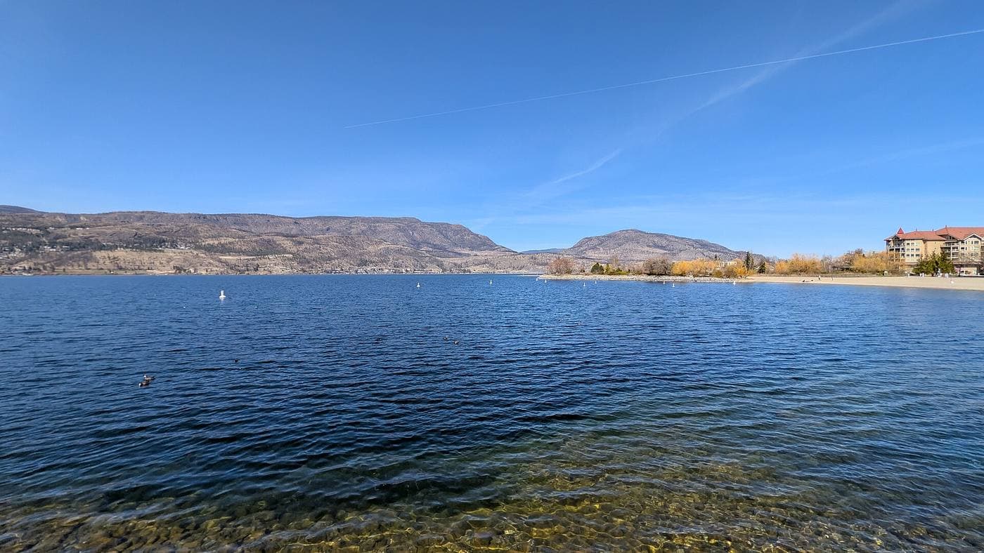 Clear shallows of Okanagan Lake over a stony bottom, with a sandy public beach and red-roofed lakeside resort at right and dry benchland hills across the water.