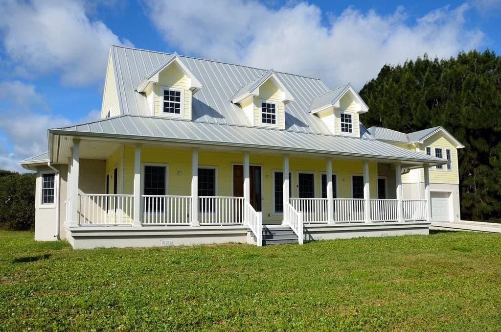 A two-storey pale yellow house with dormer windows, a silver metal roof and a white railed wraparound porch on a green lawn under blue sky.
