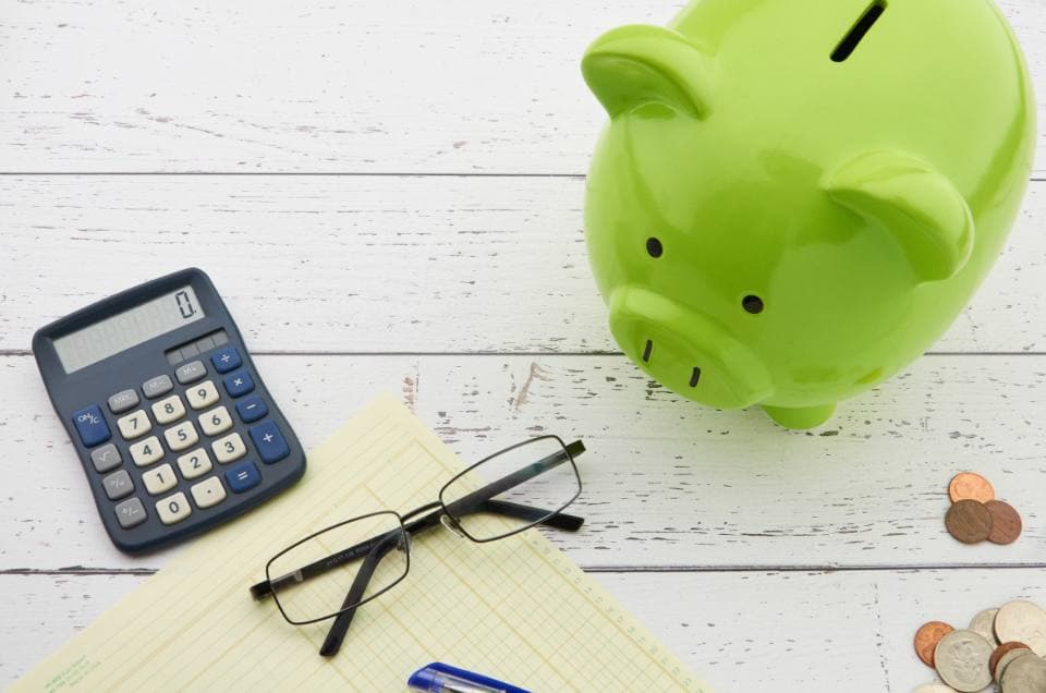 Overhead view of a green piggy bank beside a calculator, reading glasses, a ruled notepad, a pen and scattered coins on white painted wood.