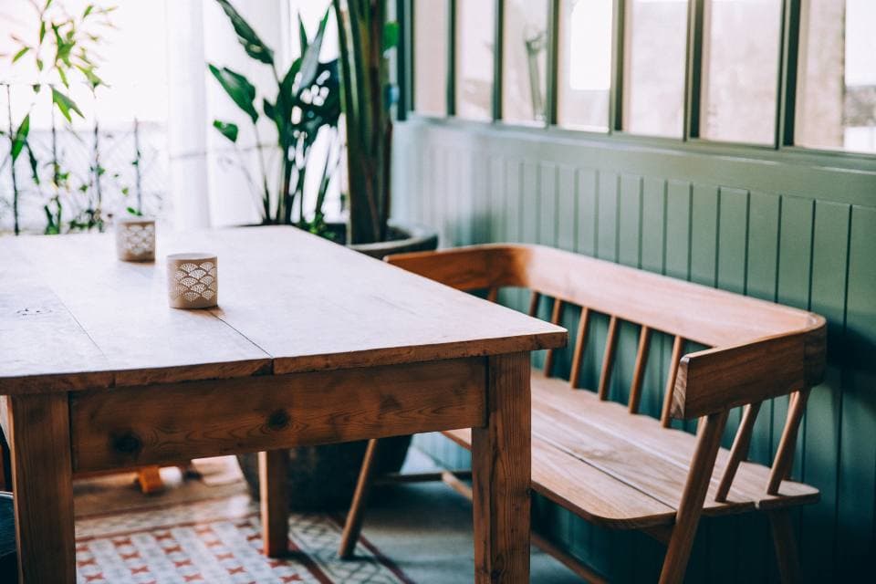 A long pine table beside a built-in wooden bench against pale green panelling, with candles, leafy plants and patterned floor tiles.