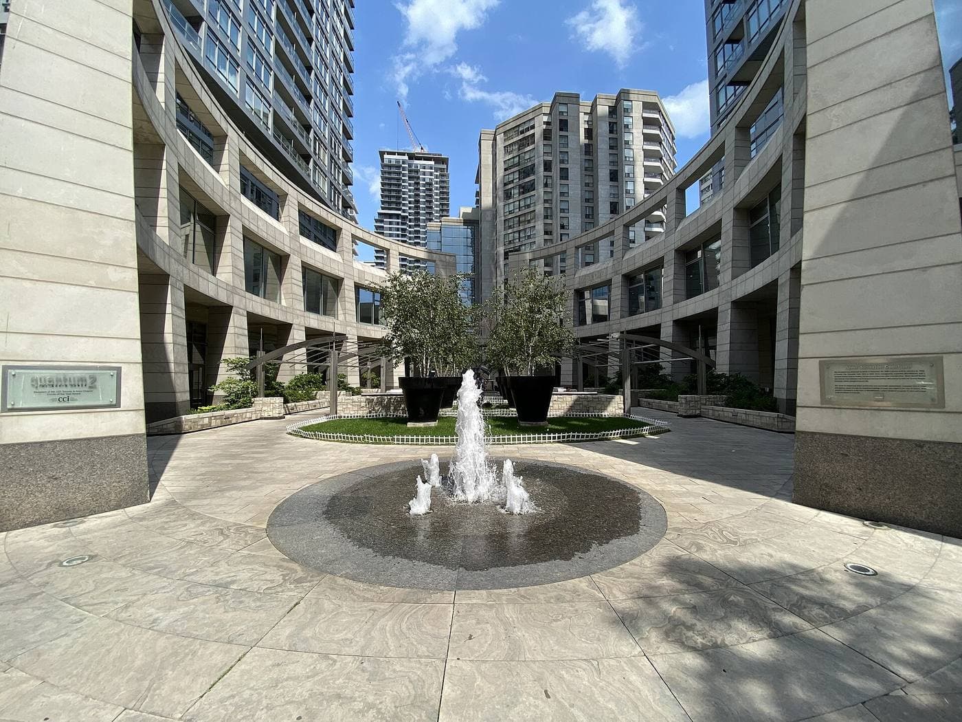 A fountain in a sunlit stone courtyard ringed by curved condominium podiums, with high-rise residential towers and a construction crane behind