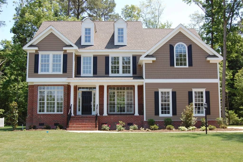 A two-storey house with shingle siding, brick base, black shutters and a white-columned front entry behind a wide mown lawn.