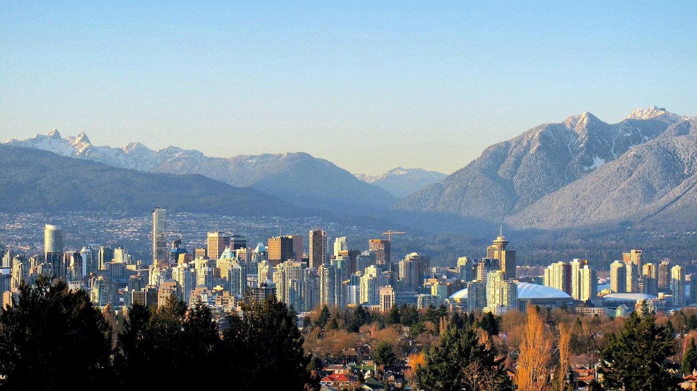 Downtown Vancouver's high-rise cluster in clear winter light, with snow-dusted North Shore mountains rising behind and treed residential streets in the foreground.