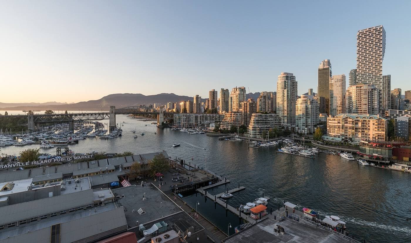 Granville Island Public Market and its marinas below False Creek condo towers, with the Burrard Street Bridge and hazy mountains in golden evening light.