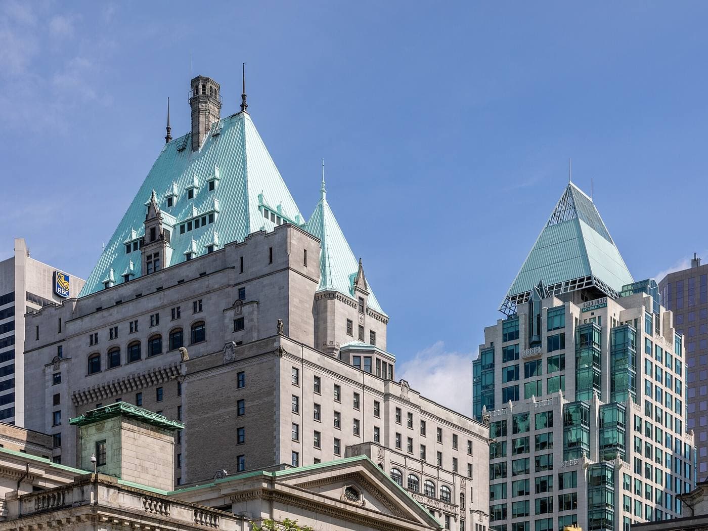 The green copper chateau roof and stone facade of the Hotel Vancouver beside the peaked glass crown of Cathedral Place under a clear blue sky.
