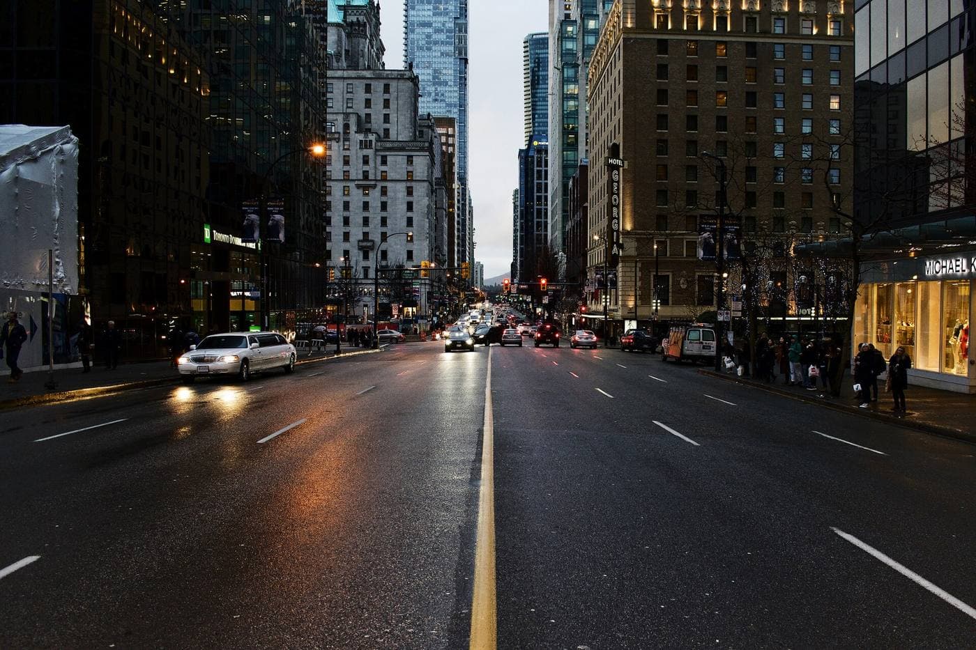 Cars and pedestrians on a wet West Georgia Street at dusk, with the Hotel Georgia and downtown office towers lining both sides of the road.