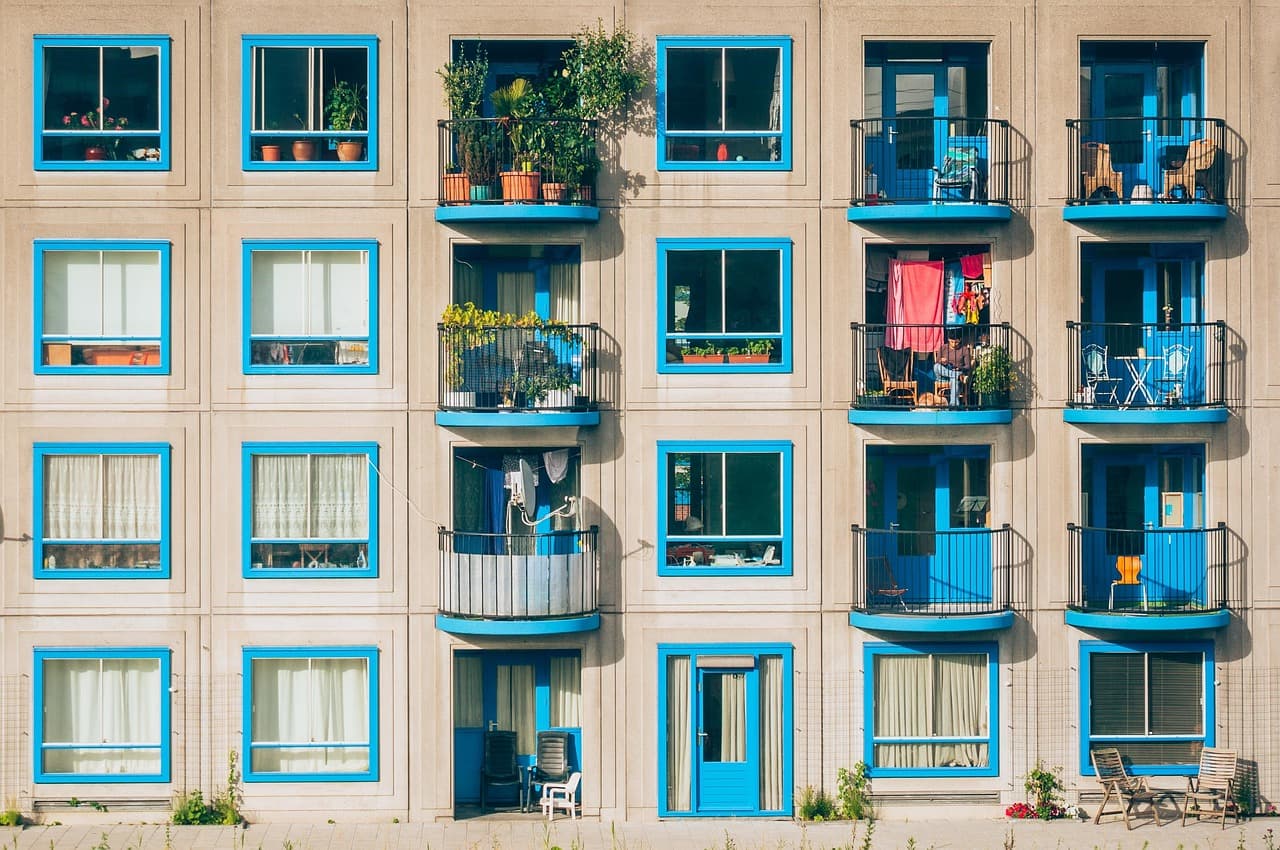 The facade of a low-rise apartment block, its balconies filled with plants and patio chairs