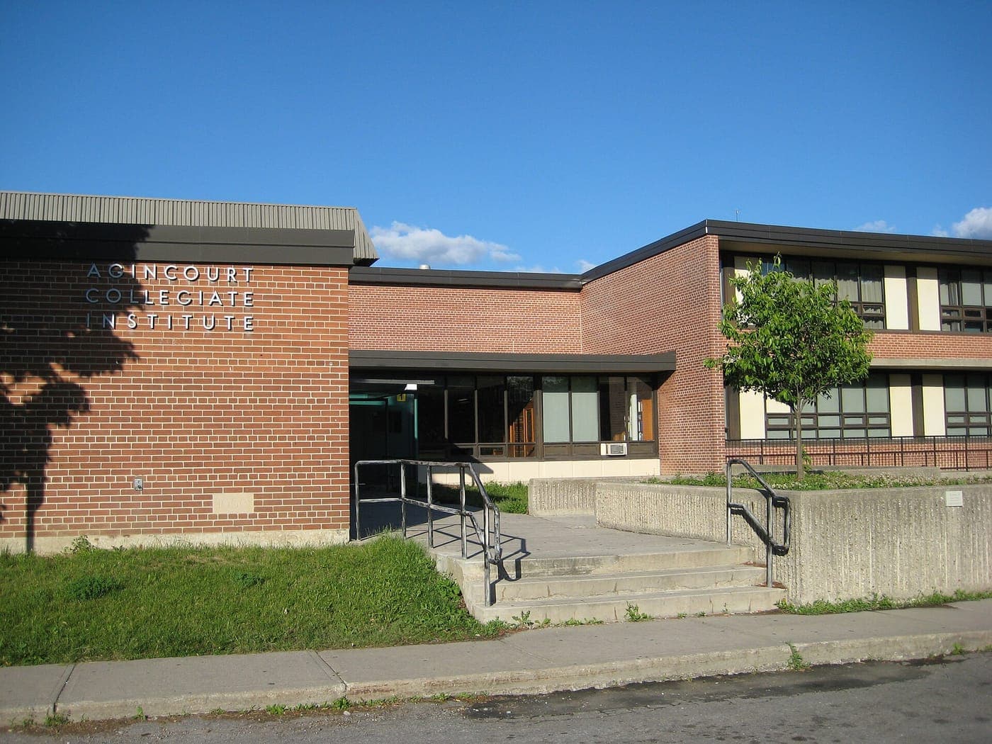 Brick facade of Agincourt Collegiate Institute with its name in metal letters, a glass entrance, concrete steps with railings and a young tree under a blue sky.