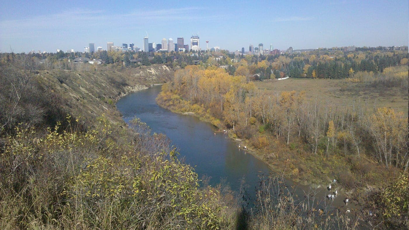 A river winds through a valley of golden autumn trees and grassy bluffs, with people and dogs along the bank and a downtown skyline on the horizon under a blue sky.