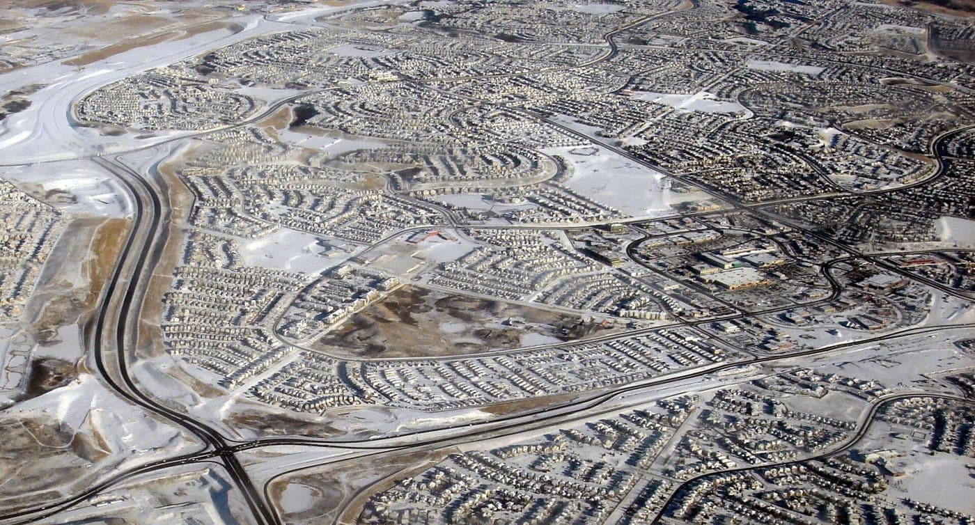 Aerial view of snow-covered suburbs with curving streets lined with detached houses, a divided highway along the left edge, open green spaces and a shopping area on the right.