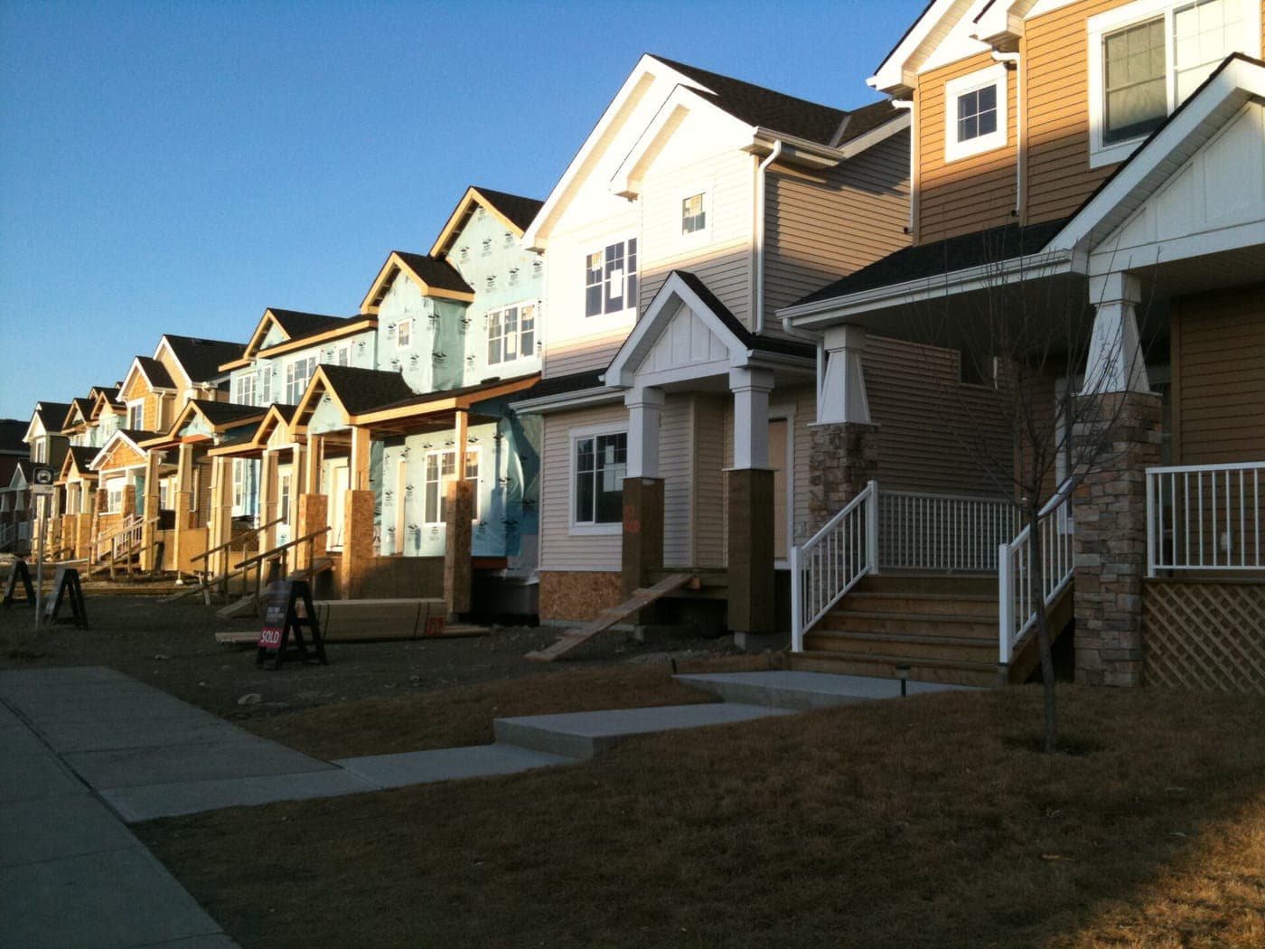 A row of two-storey houses with covered front porches along a sidewalk, finished homes in front and more still wrapped and under construction beyond, under a clear blue sky.