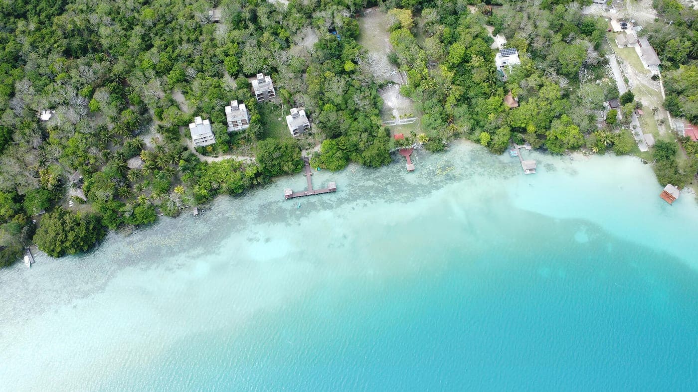 Aerial view of a jungle-lined lagoon shore with white shallows fading into turquoise water, wooden docks and several white houses among palm trees.