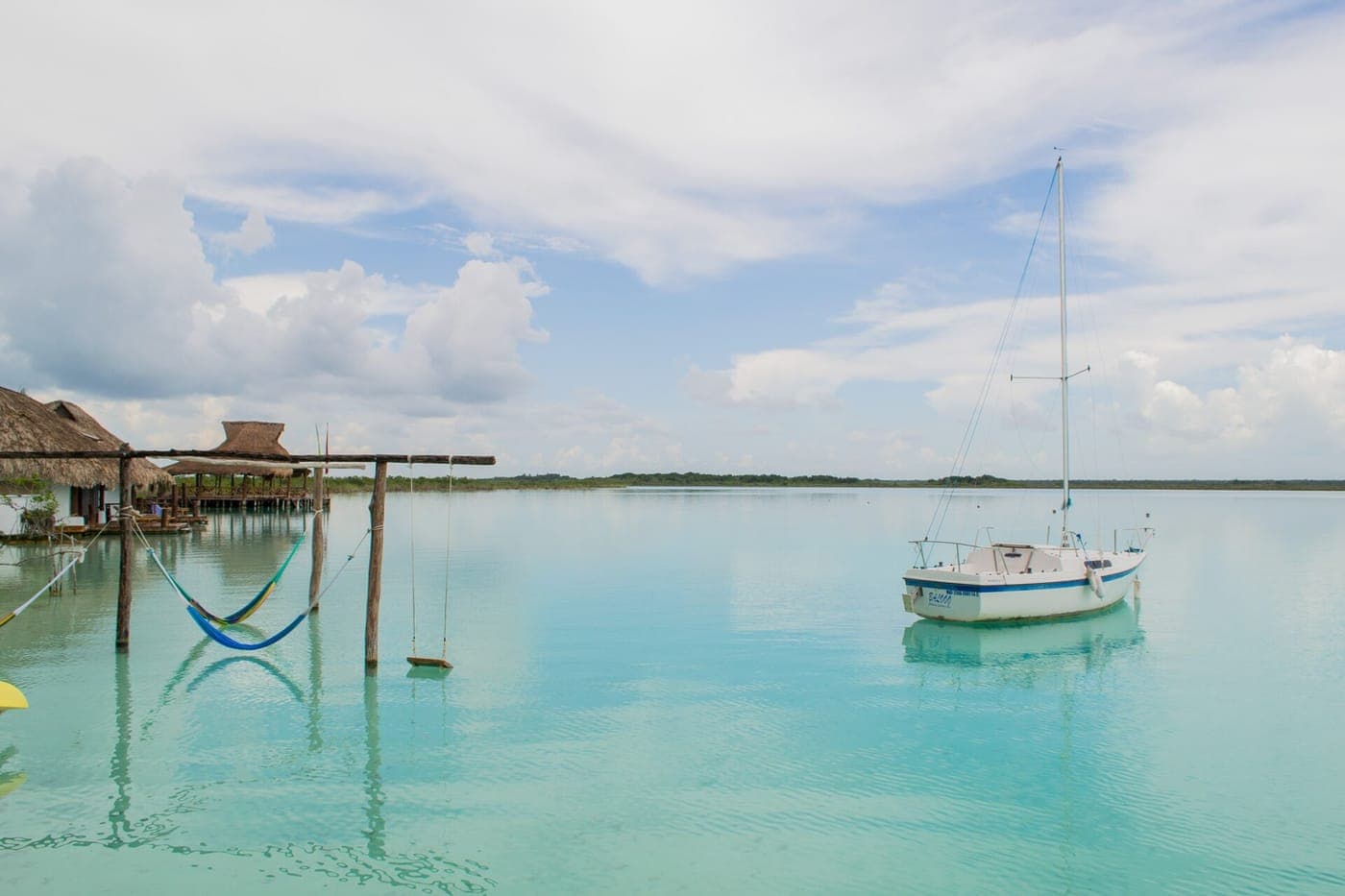 Hammocks and a wooden swing hang from posts over calm turquoise lagoon water beside thatched-roof palapas, with a white sailboat moored nearby under a cloudy sky.