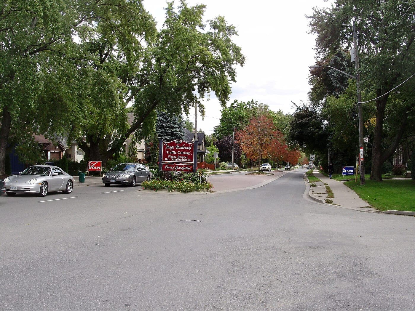 Tree-lined residential street with a red traffic-calming sign on a landscaped median, parked cars, houses behind mature trees and early autumn foliage