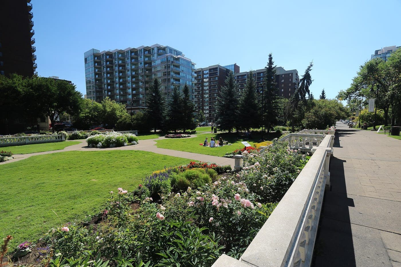Formal park gardens with flower beds, lawns and white stone balustrades beside a paved walkway, with mid-rise and high-rise apartment buildings and evergreen trees behind under a clear blue sky.