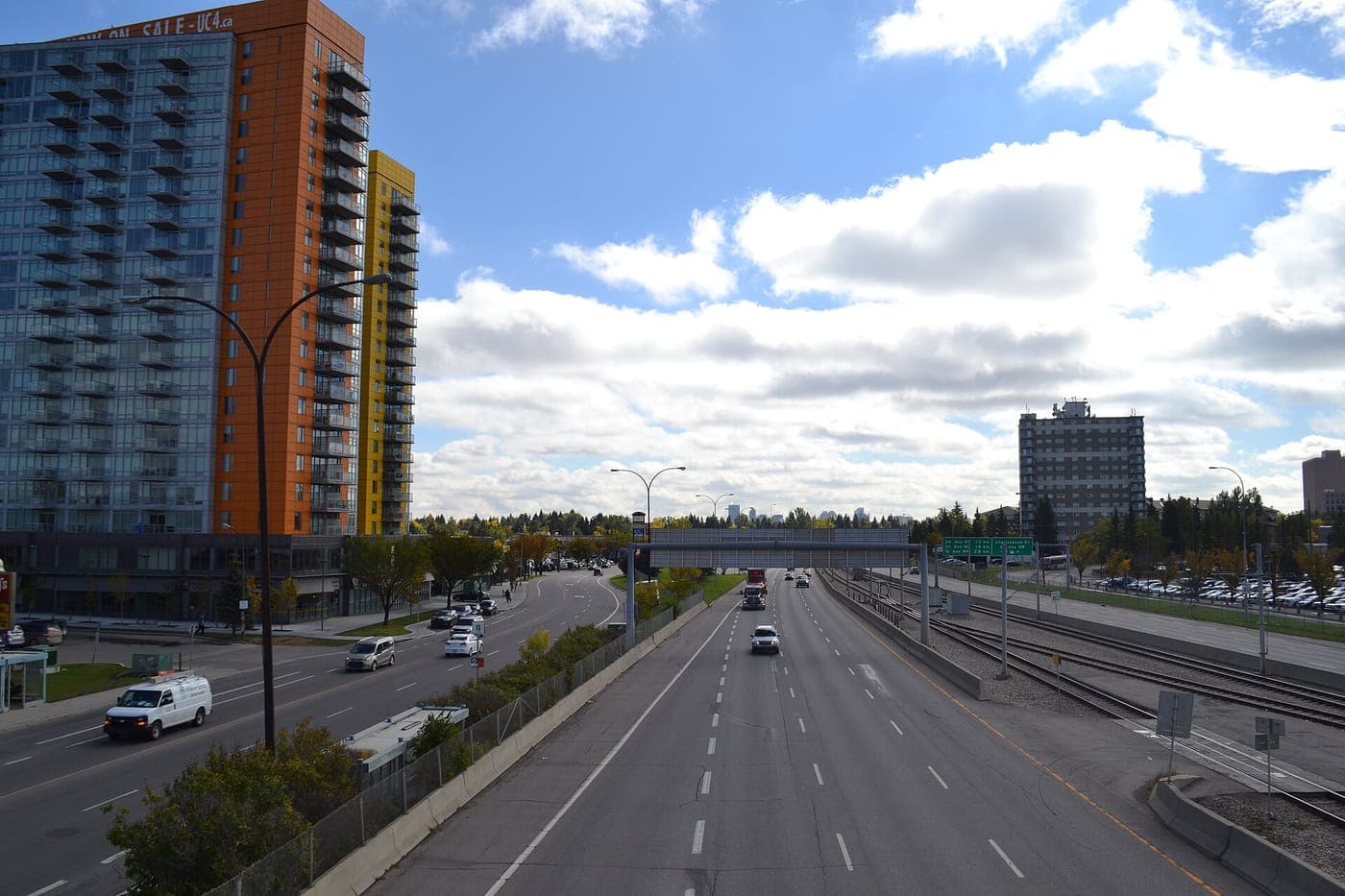 Colourful high-rise apartment towers beside a multi-lane street and a divided highway with light rail tracks, with trees and a mid-rise building under a partly cloudy sky.