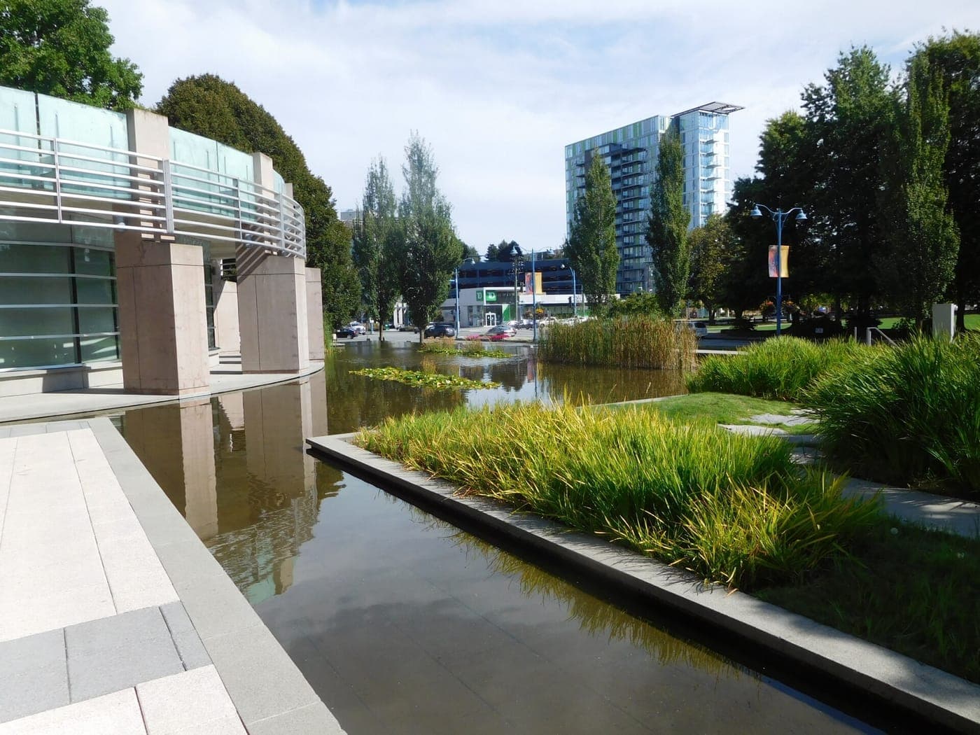 Landscaped reflecting pools with tall grasses beside a curved glass-and-stone building, with trees, a street, and a high-rise apartment tower in the background on a sunny day.