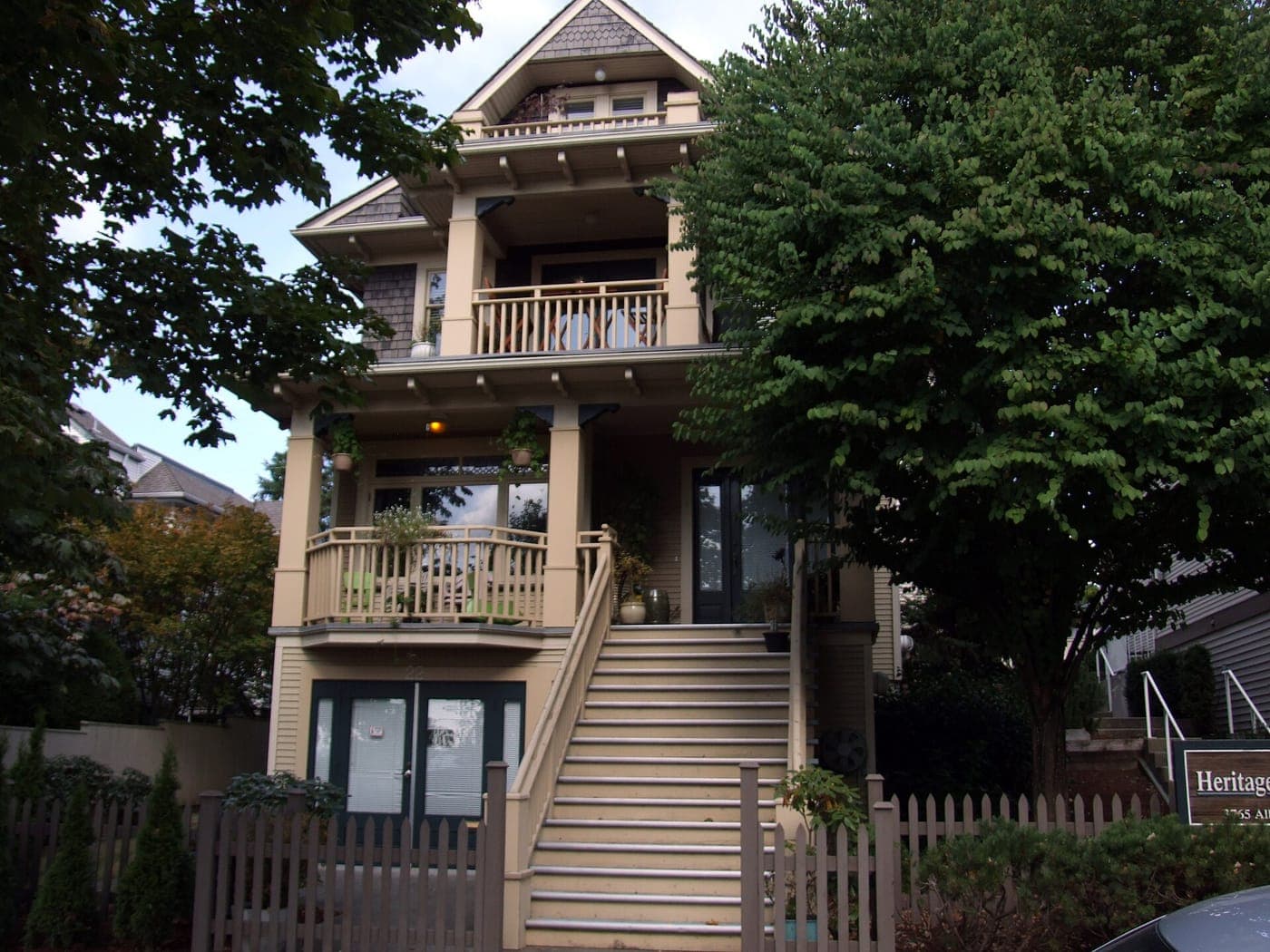 Three-storey Craftsman-style house with stacked front balconies, a tall front staircase and picket fence, framed by large leafy trees