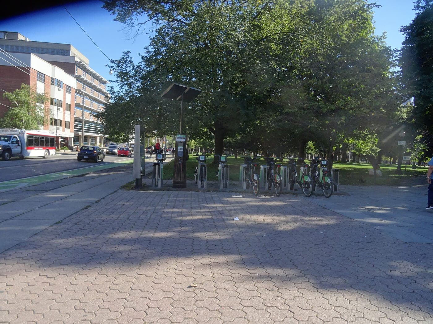 A bike share station with green bicycles docked on a brick-paved plaza beside a tree-filled park, with a street, passing traffic, a shuttle bus and mid-rise buildings on the left.