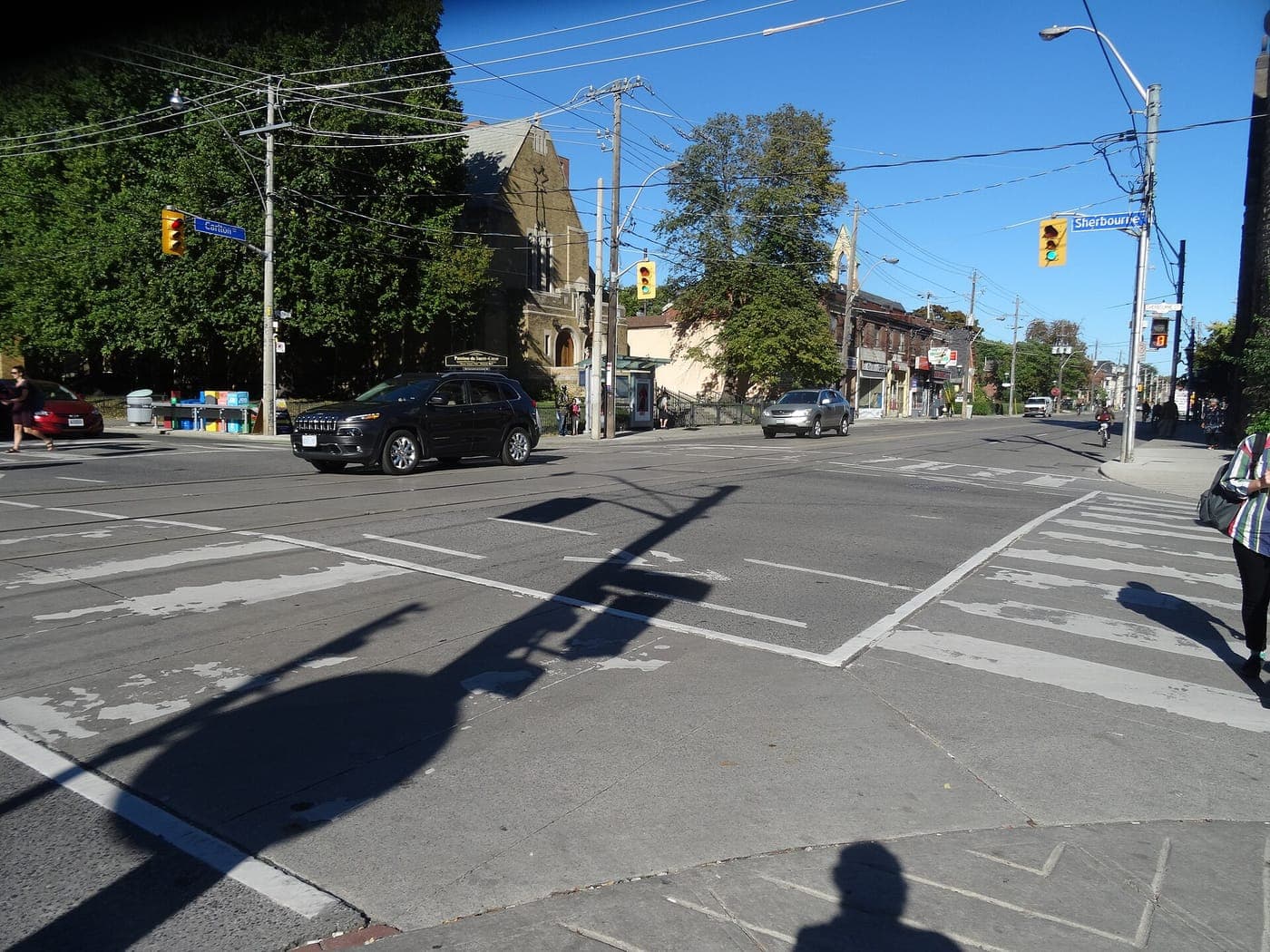 Sunny urban intersection with streetcar tracks, crosswalks and traffic lights, a stone church among trees, cars, and a row of low-rise shops and houses lining the street beyond