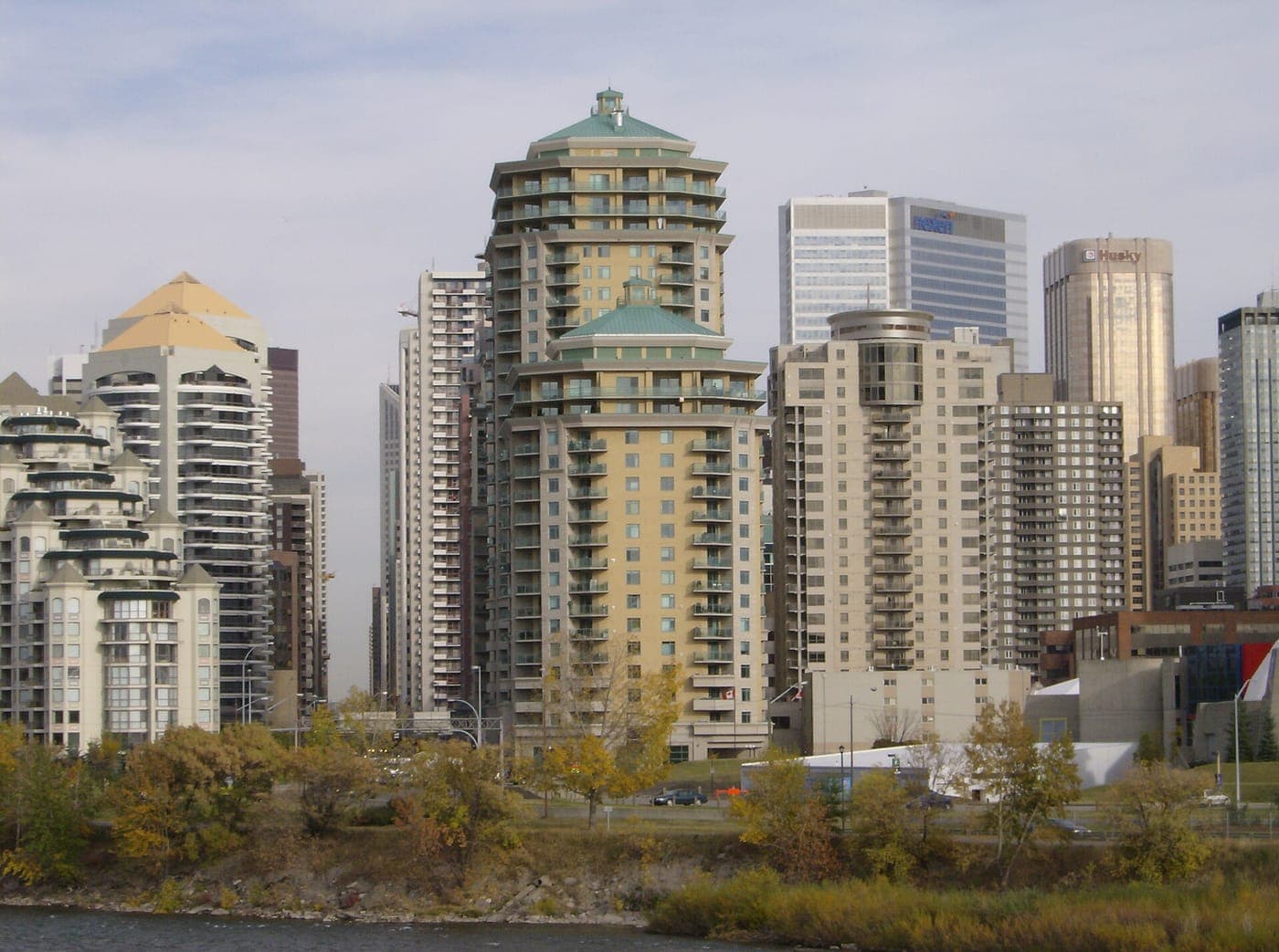 Residential high-rise towers with green and gold pyramid roofs stand above a riverbank lined with autumn trees, with downtown office towers rising behind them.