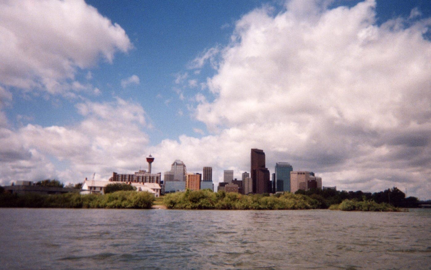 Downtown skyline with a tall observation tower and high-rise office buildings rising behind green shrubs along a river under a blue sky with large white clouds.
