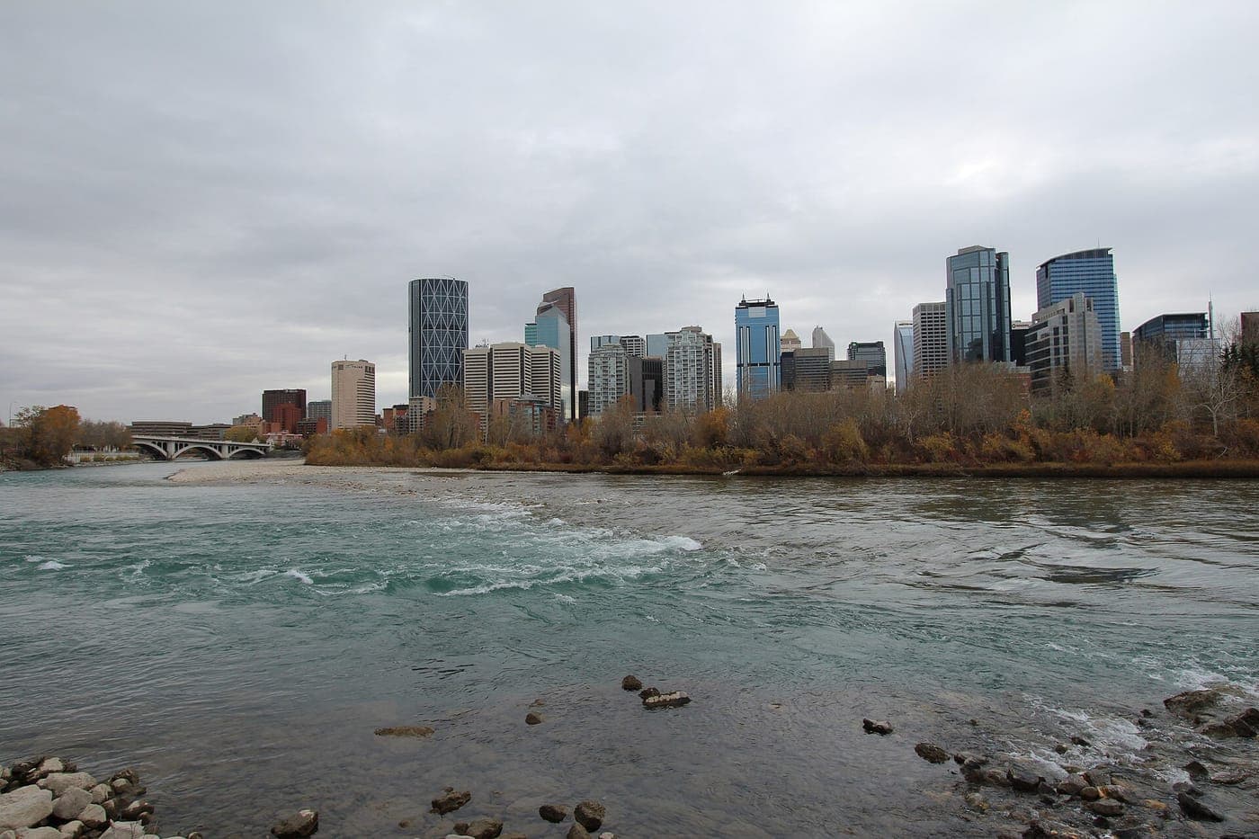 Calgary's downtown skyline of glass high-rises, including a diagrid-patterned tower, rises beyond the teal Bow River with a rocky shore, an arched bridge and autumn trees under grey skies.