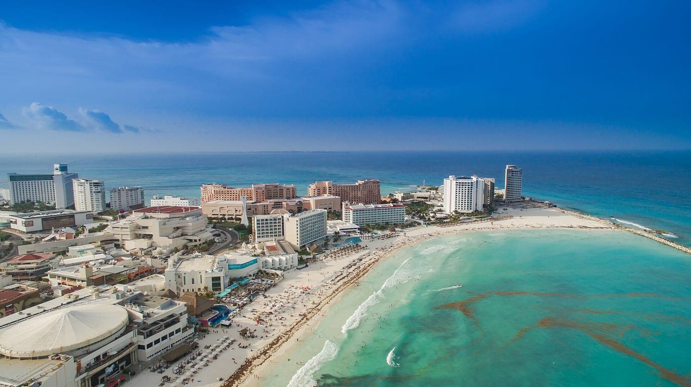 Aerial view of high-rise beachfront hotels and resorts along a curving white-sand beach with turquoise Caribbean water at Punta Cancún in the Hotel Zone