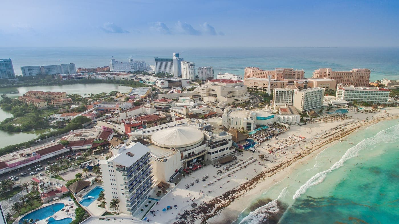 Aerial view of Cancún's Hotel Zone, with high-rise resorts, pools and a white domed building lining a sandy beach between a green lagoon and the turquoise Caribbean Sea.