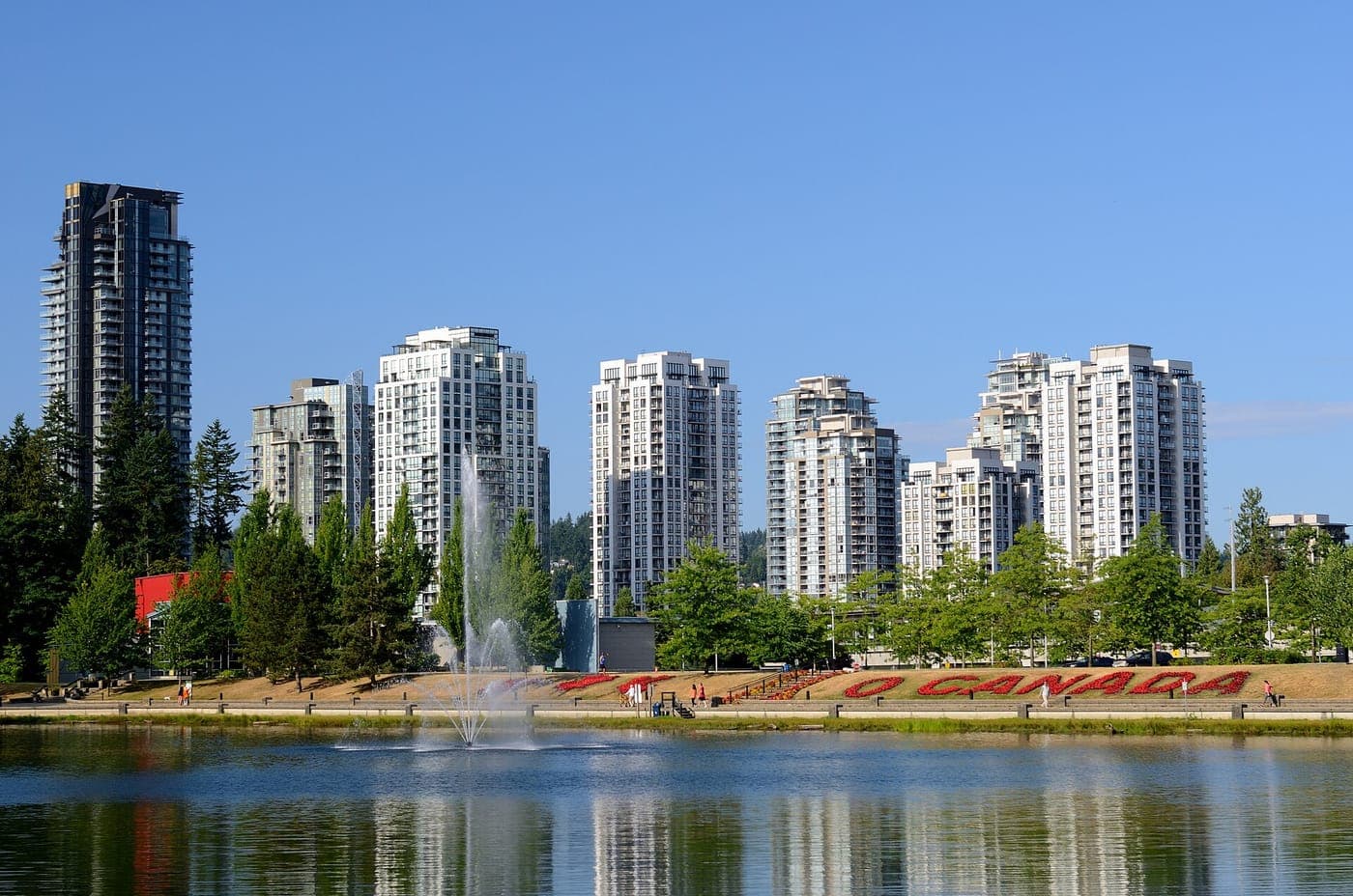 High-rise condo towers stand behind a calm lake with a fountain and a lawn where red flowers spell O CANADA, under a clear blue sky.