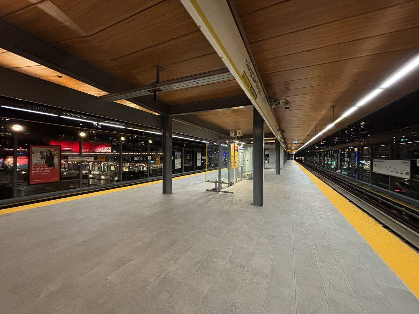 Empty elevated transit platform at night with a warm wooden slatted ceiling, yellow tactile edge strip, glass shelter and lit city buildings beyond the tracks.