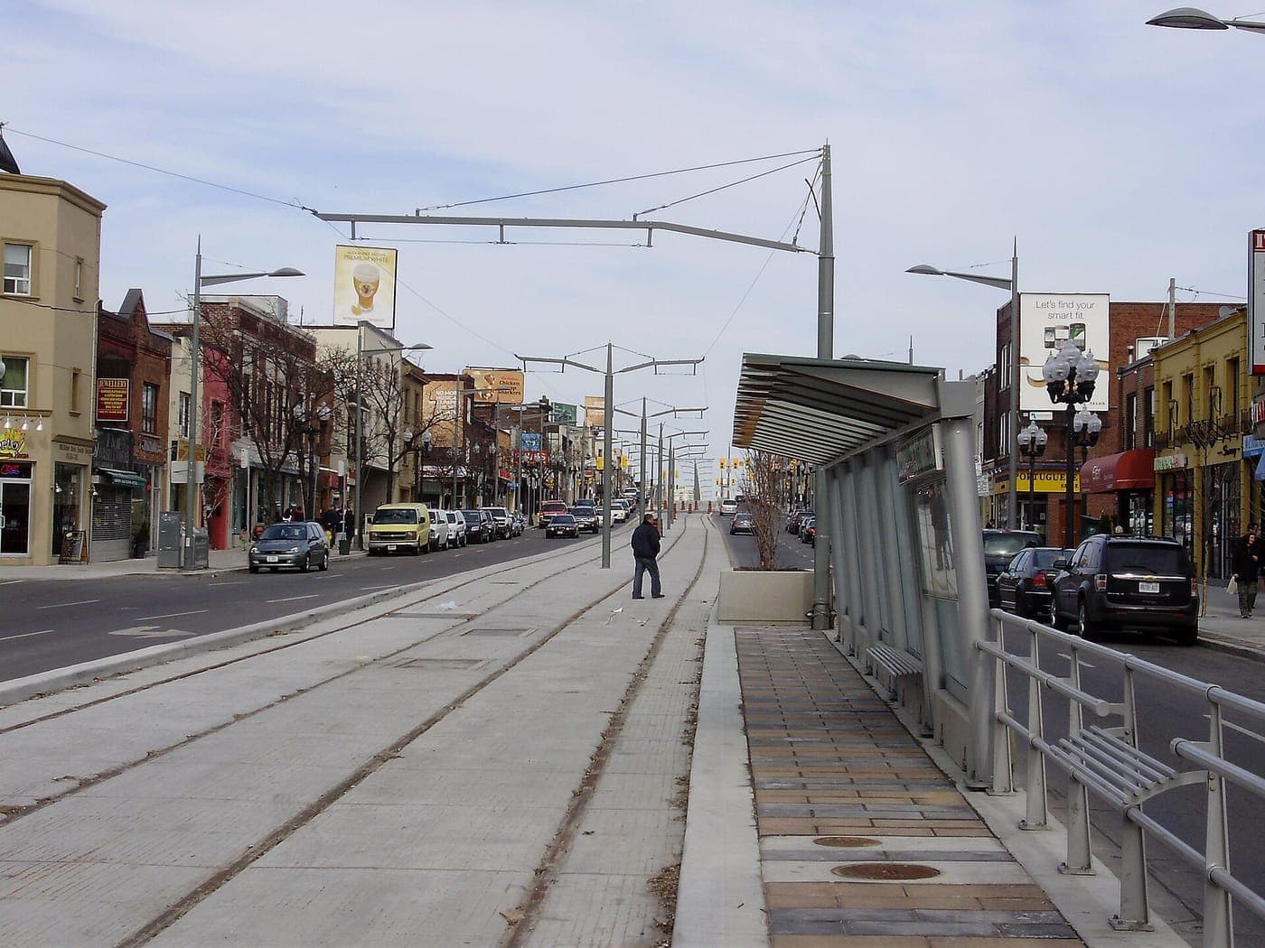 A city avenue with a streetcar right-of-way and a covered transit platform down the middle, lined on both sides by low-rise shops, parked cars, ornate street lamps and overhead wires.