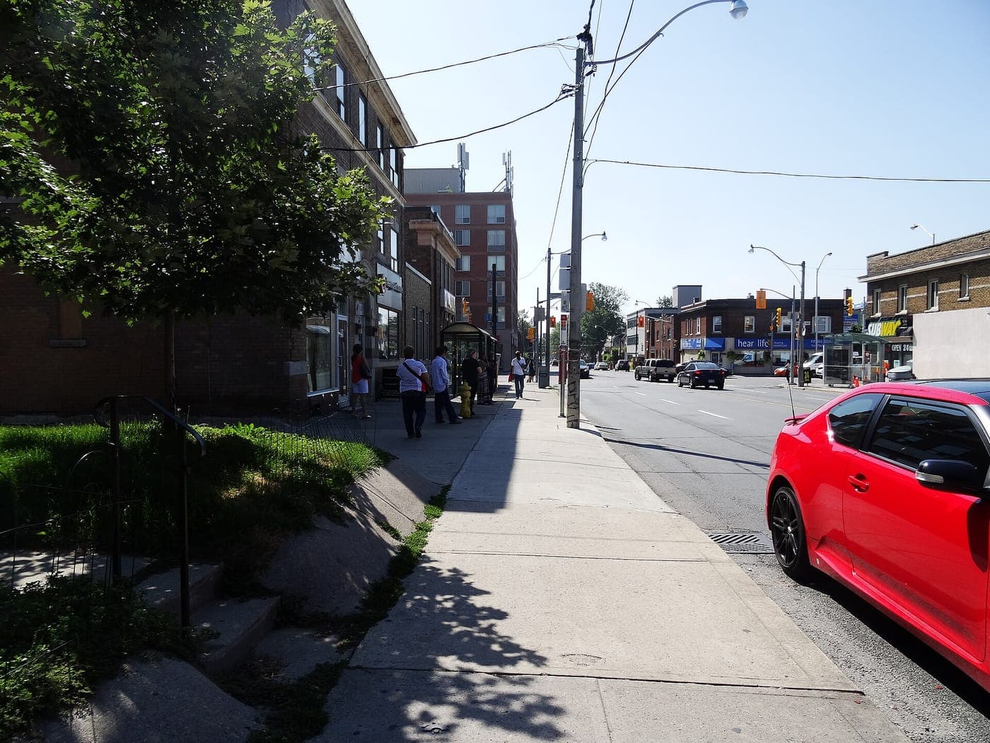 Sunny sidewalk along a main street lined with low-rise brick buildings, a bus shelter with people waiting, utility poles and shops, and a red car parked at the curb.