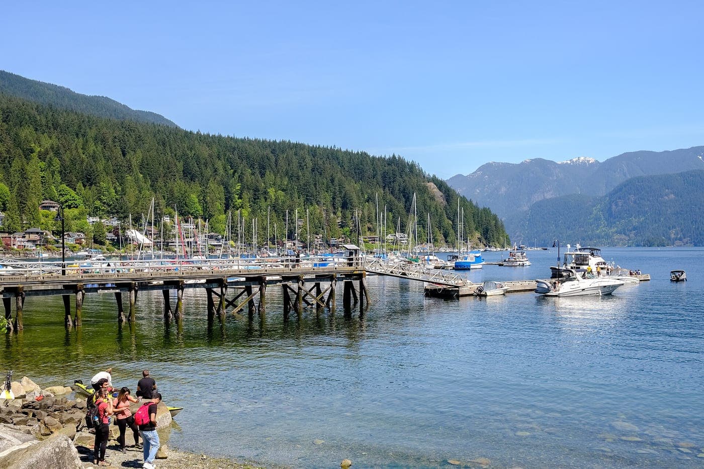 Wooden pier and marina full of sailboats in Deep Cove, with forested hillside homes, calm blue water, distant snow-capped mountains and a few people on the rocky shore