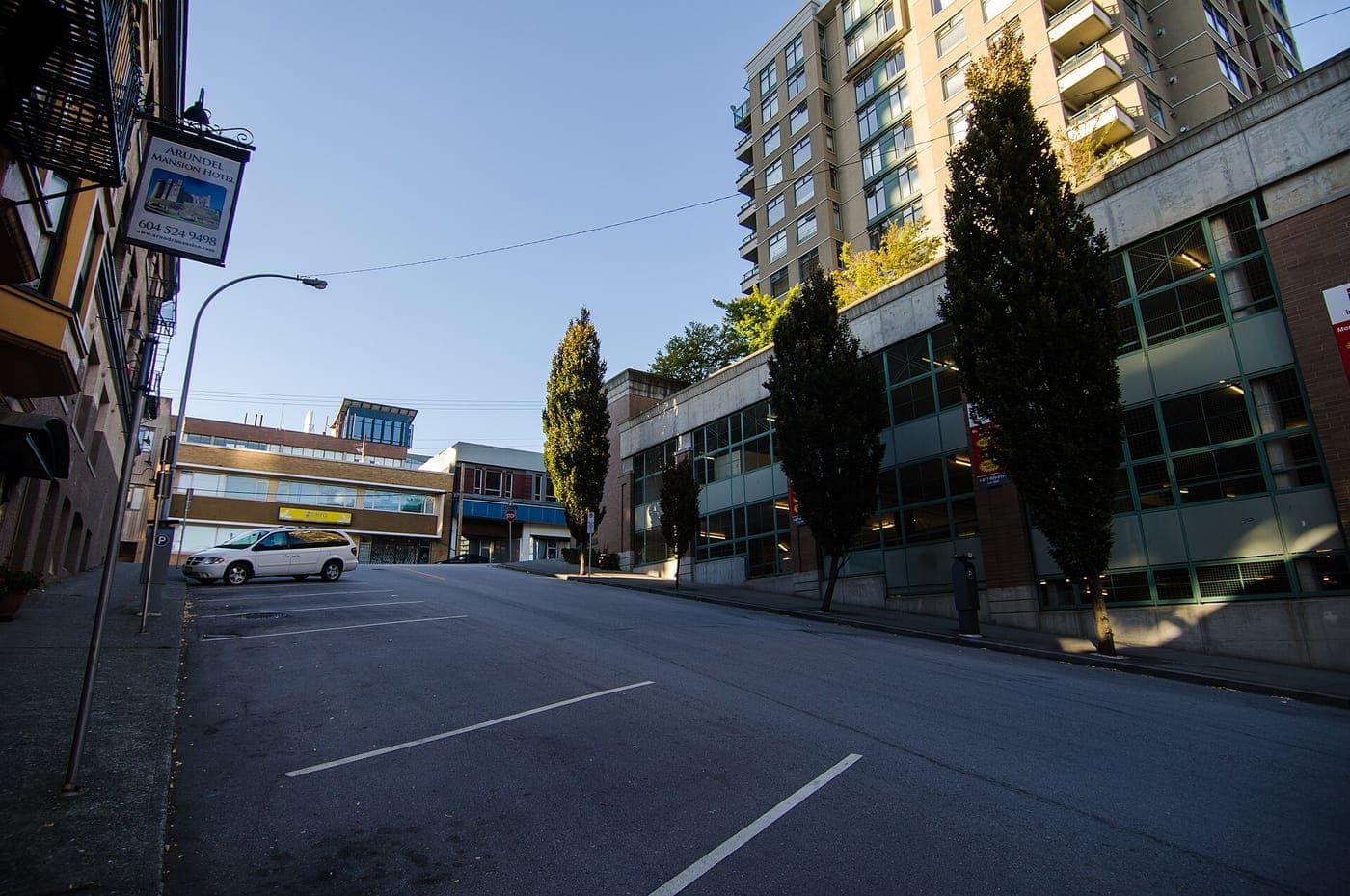 Sloping urban street with an empty parking area, a parked white minivan, tall columnar trees along a low glass-fronted building, and a high-rise condo tower under a clear blue sky