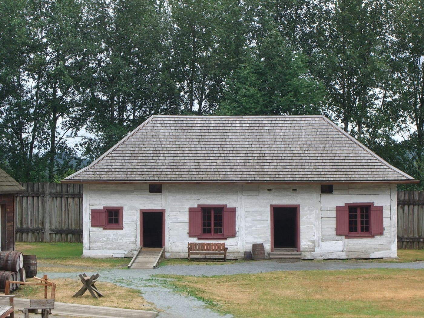 Whitewashed log building with red shutters and doors and a hipped cedar shake roof, set inside a wooden palisade with barrels, a bench, a gravel path and tall trees behind
