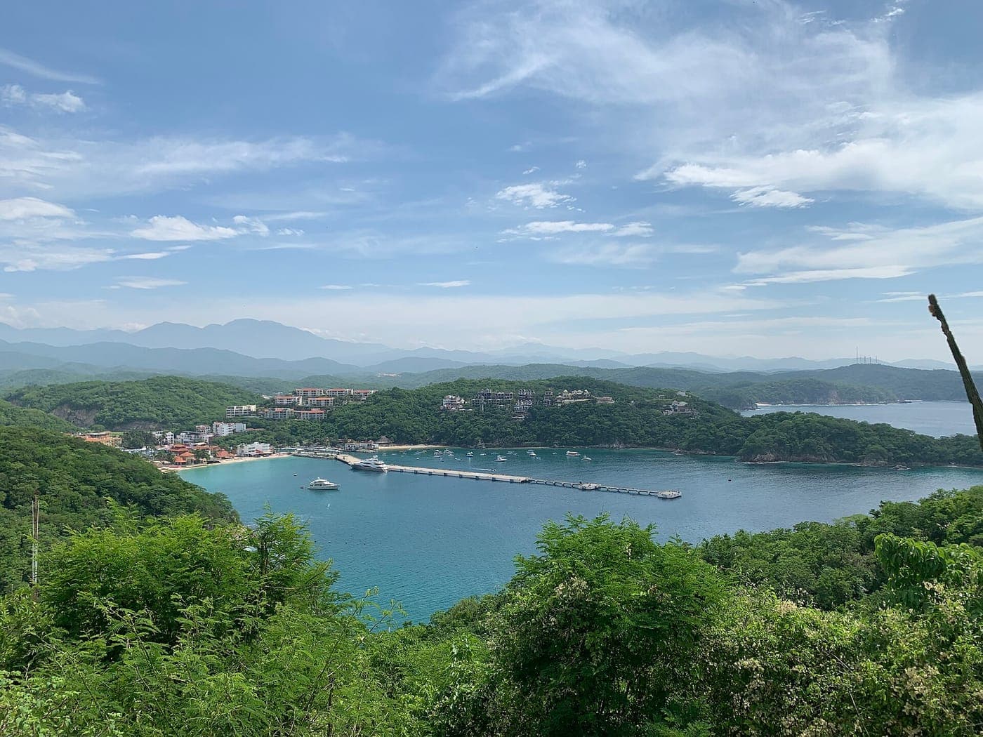Elevated view over a turquoise bay with a long pier, moored boats, a sandy beach and hillside resort buildings surrounded by green forest, with mountains under a blue sky.