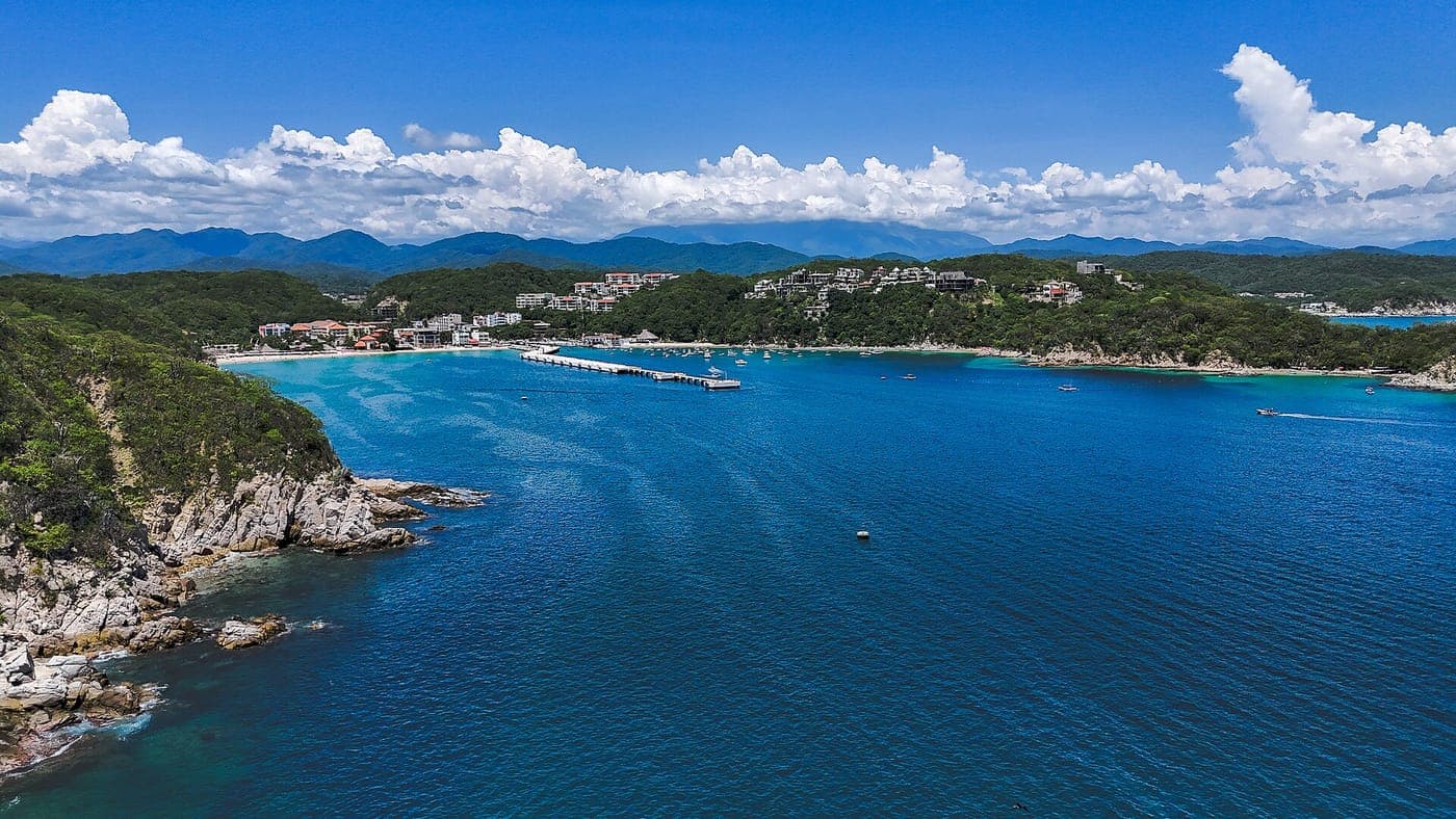 Aerial view of a deep blue bay with a long pier, small boats, a sandy beach and hillside buildings among green forest, with rocky shoreline in front and mountains under white clouds