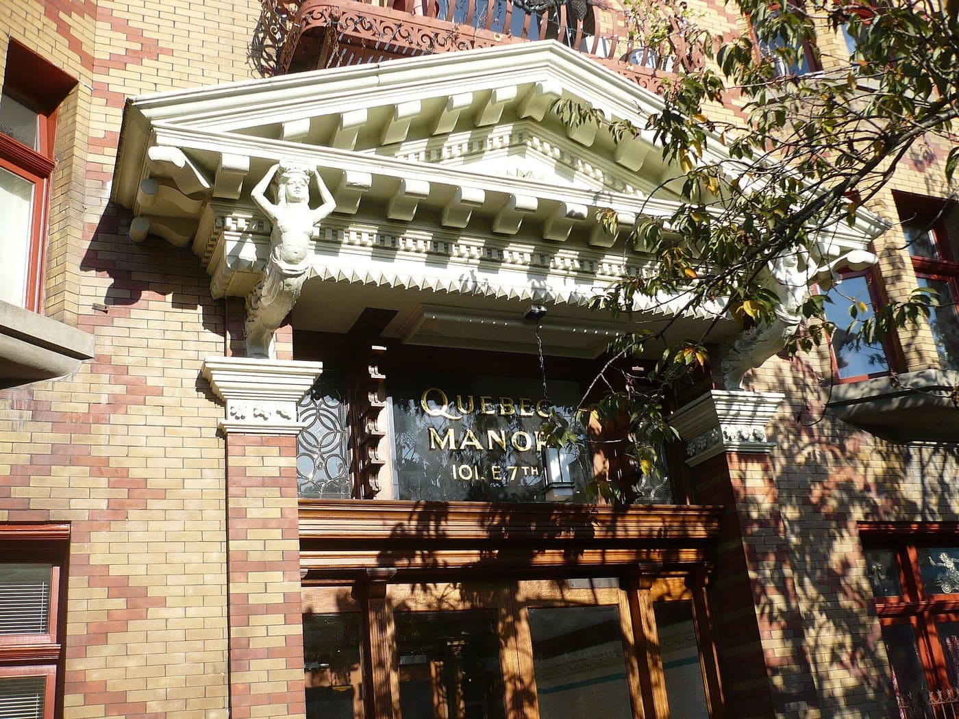 Ornate white pediment with a carved caryatid figure over the wood-framed entrance of a patterned brick heritage apartment building, with gold lettering reading Quebec Manor 101 E 7th