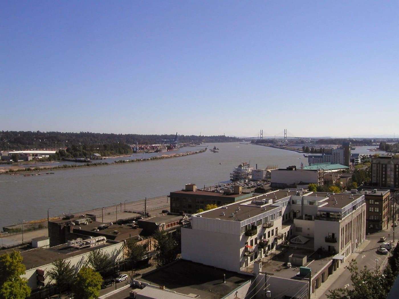 Elevated view over low-rise downtown buildings and a riverside street toward a wide river, with docks, a moored riverboat and a cable-stayed bridge in the distance under clear blue sky.