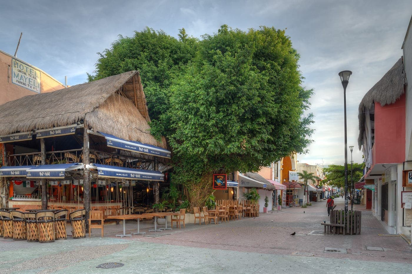 Quiet pedestrian street with a thatched palapa-roof restaurant, outdoor wooden tables and chairs, a large leafy tree, pastel shopfronts and a tall streetlamp under a cloudy sky
